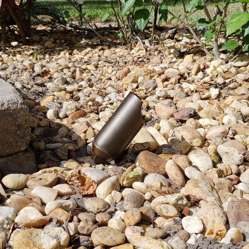 Brown cylindrical landscape spotlight lies among light-colored stones in a garden bed.
