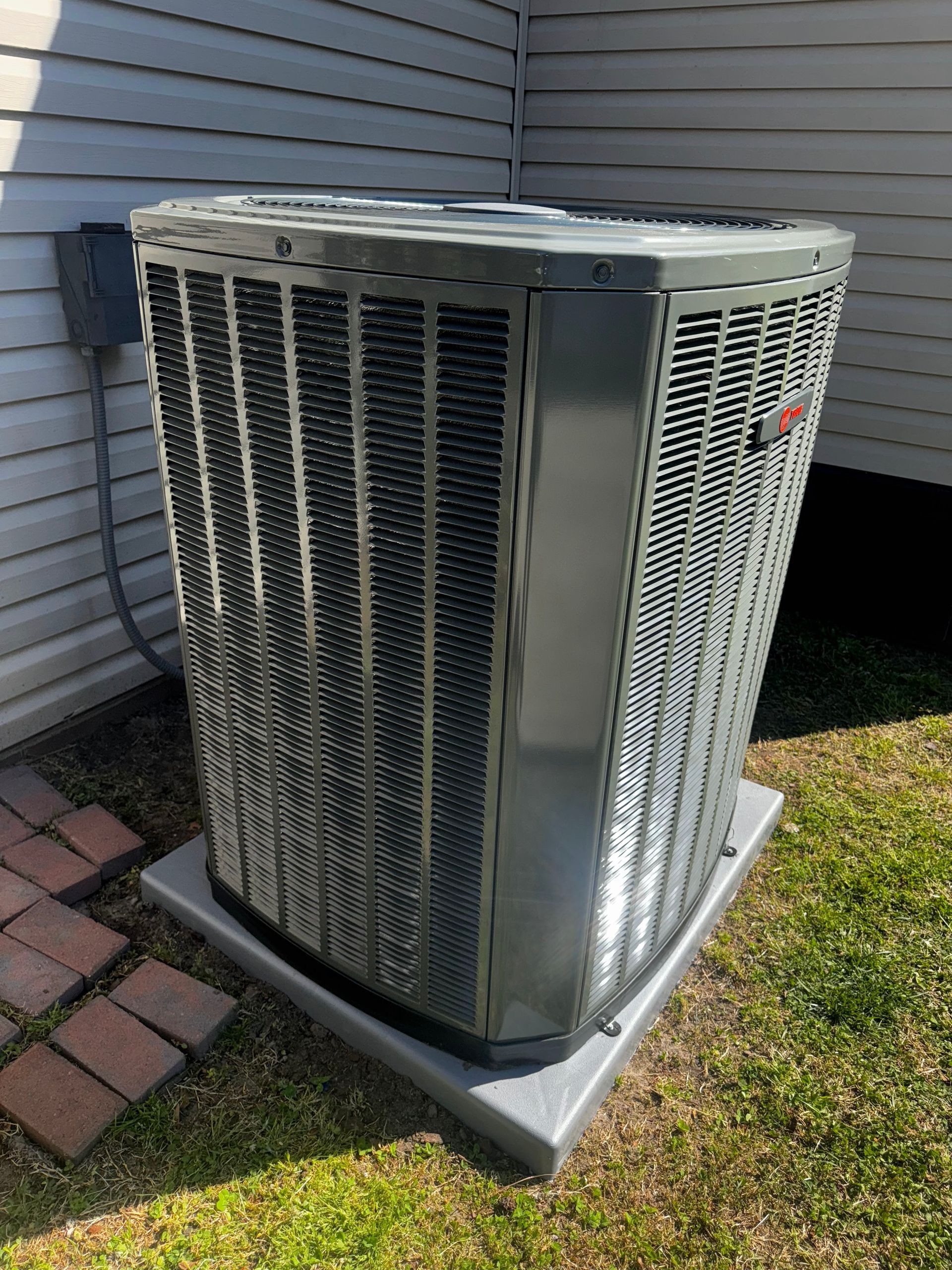 Gray air conditioning unit outside on a concrete pad next to a house and brick walkway.