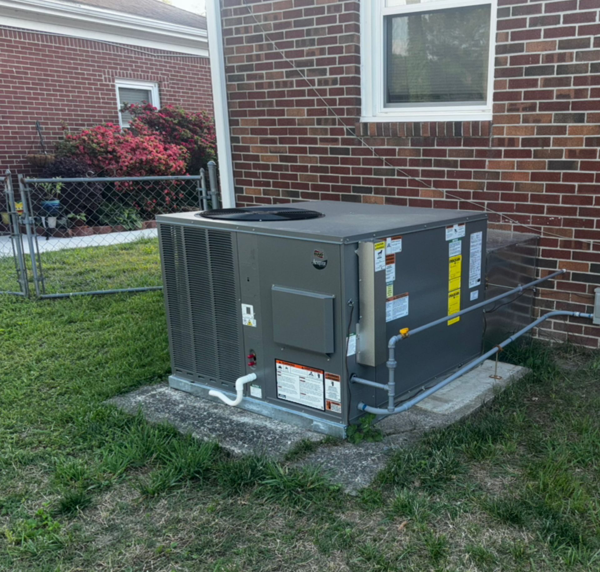 Gray air conditioning unit outside a brick building on a concrete pad, next to grass and a chain link fence.