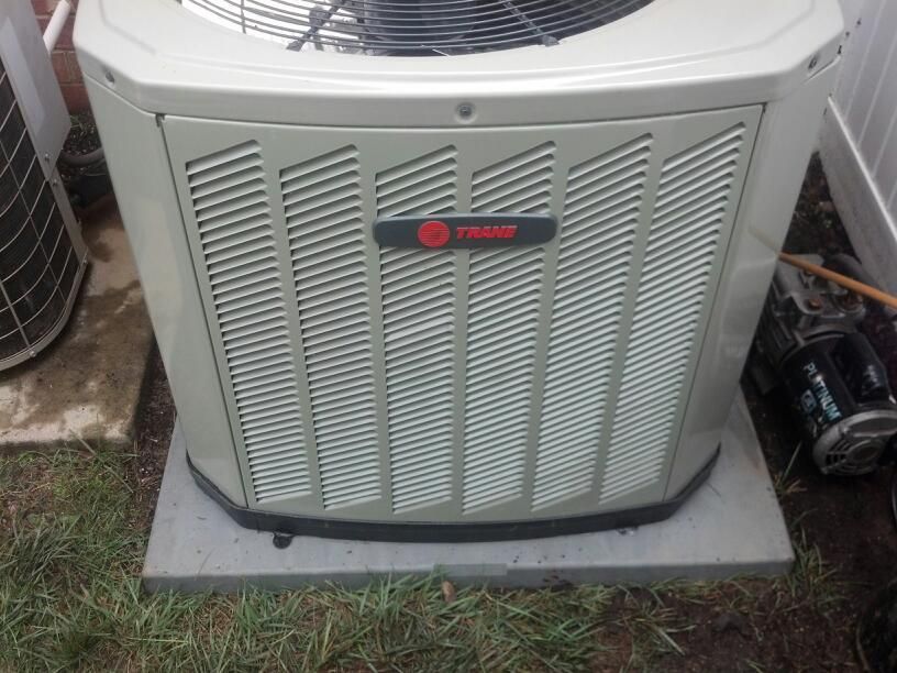 Trane air conditioning unit on a concrete pad, surrounded by grass, with a white fence visible.