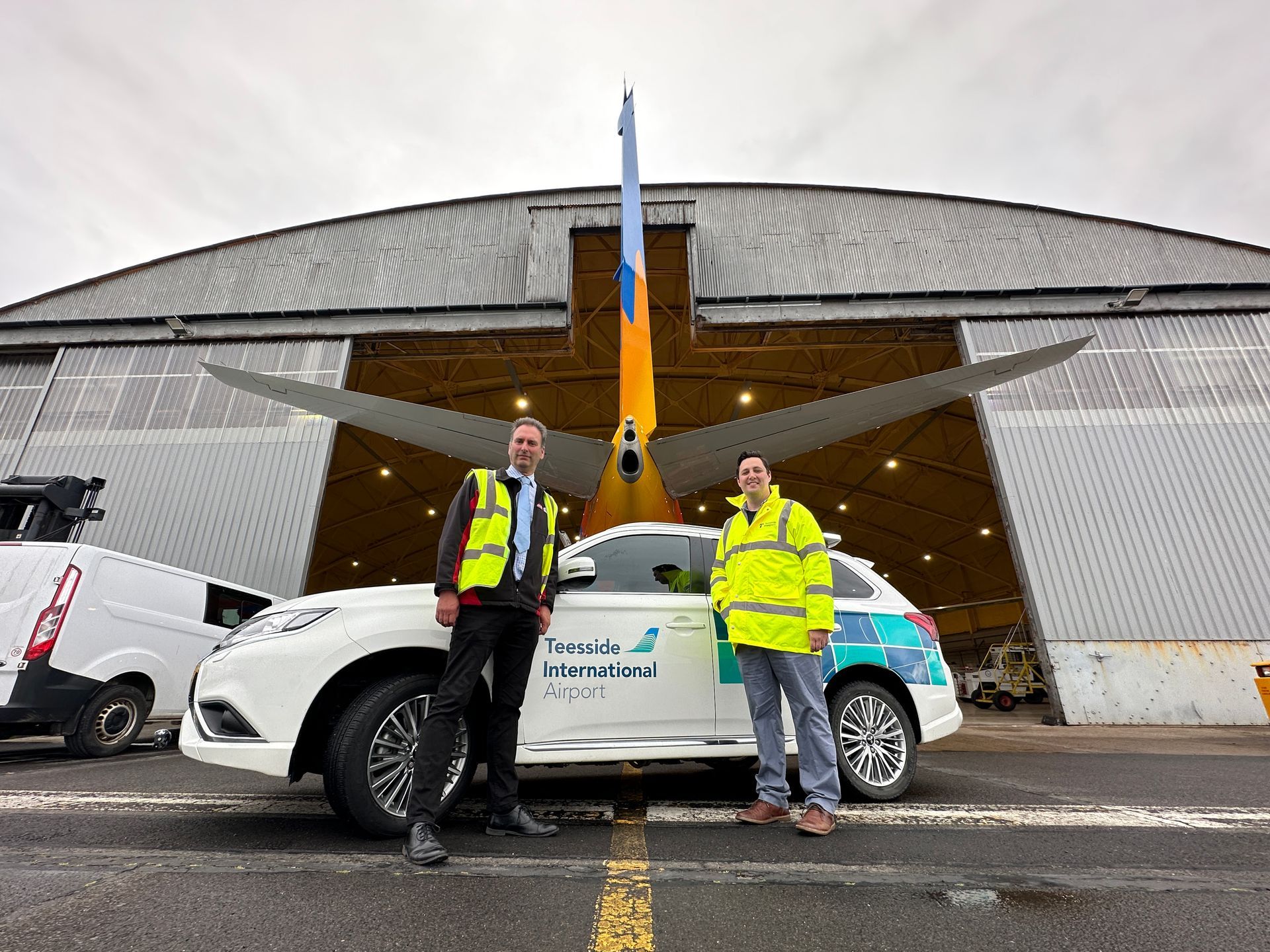 Two men are standing next to a car in front of an airplane hangar.