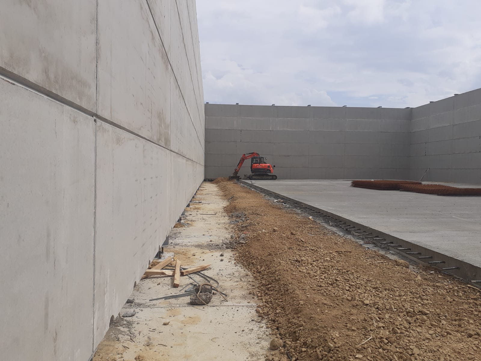 A large concrete wall is being built with a red excavator in the background.