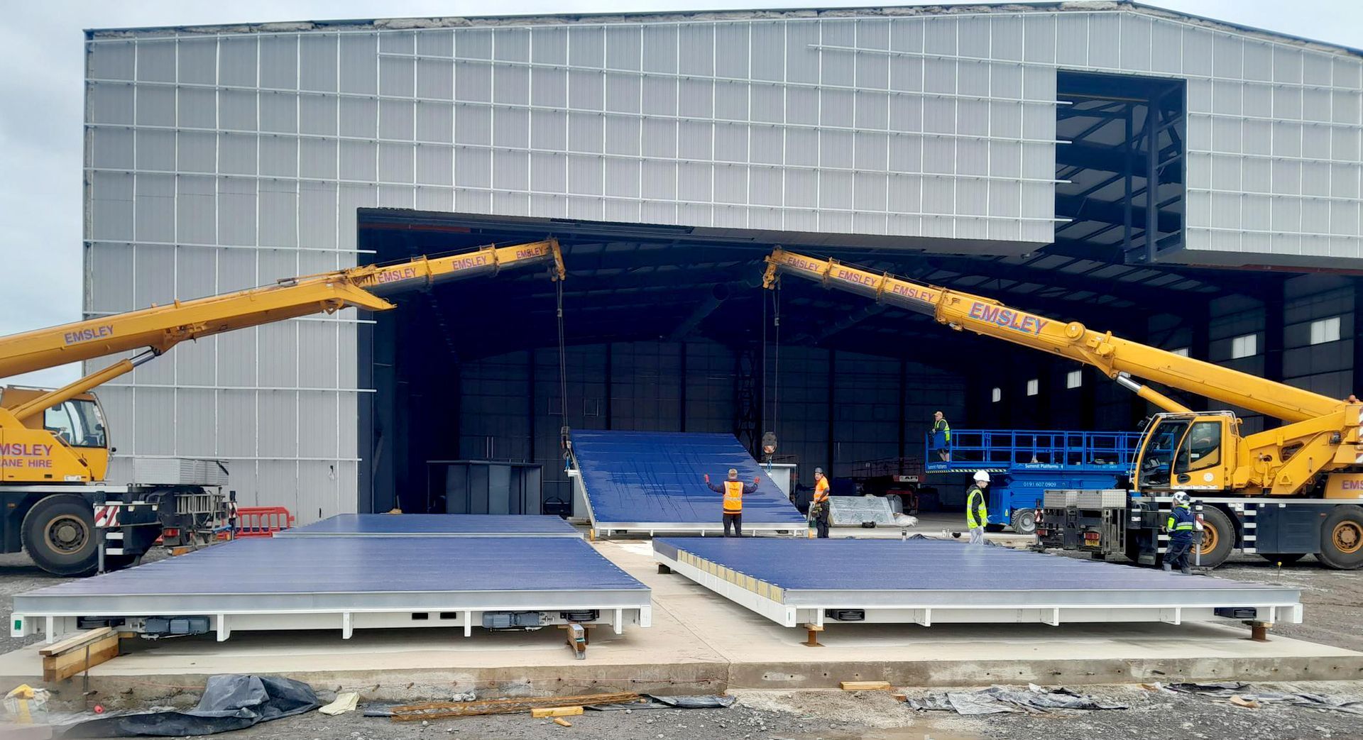 Two yellow cranes are lifting a large piece of metal in front of a large building.