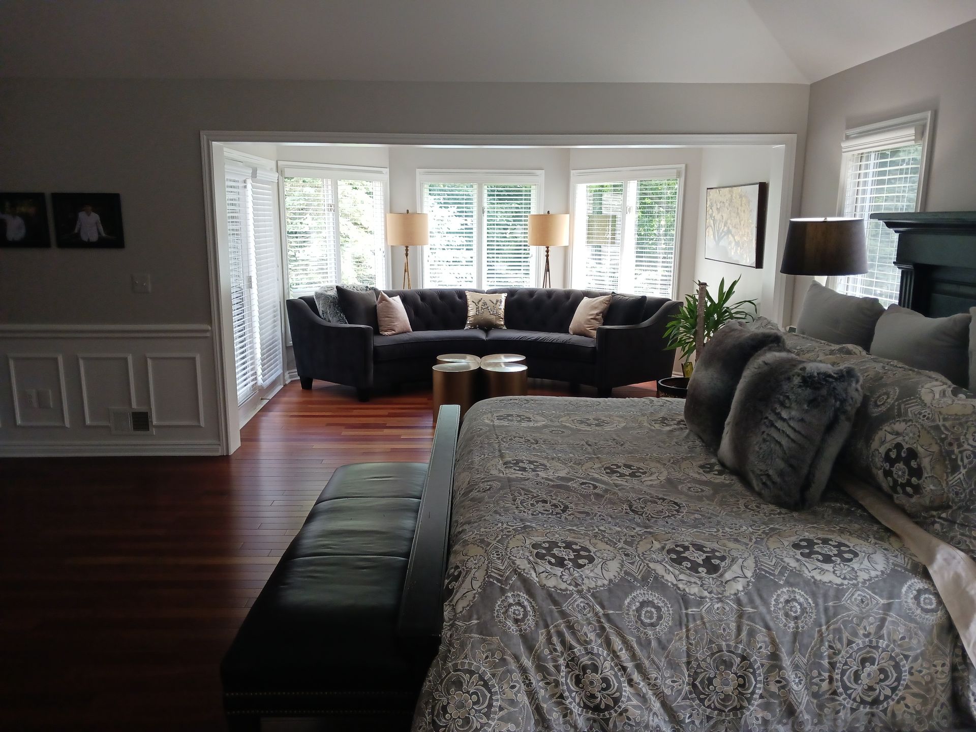 Spacious bedroom with dark sofa, large windows, dark wood floors, and a king-sized bed. 