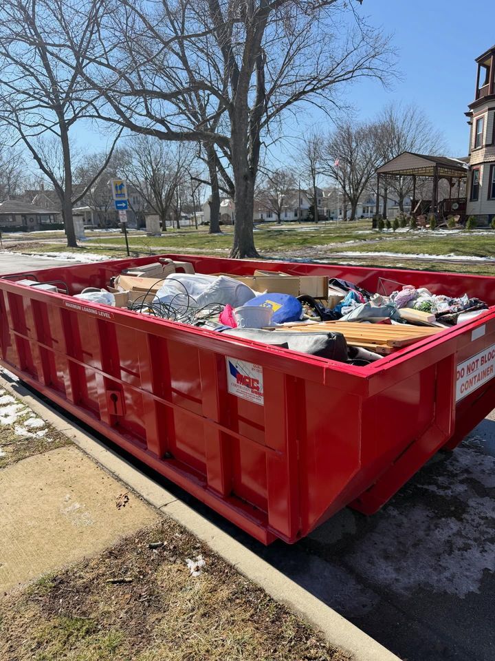 Large red construction dumpster filled with debris beside a sidewalk and leafless trees on a sunny day