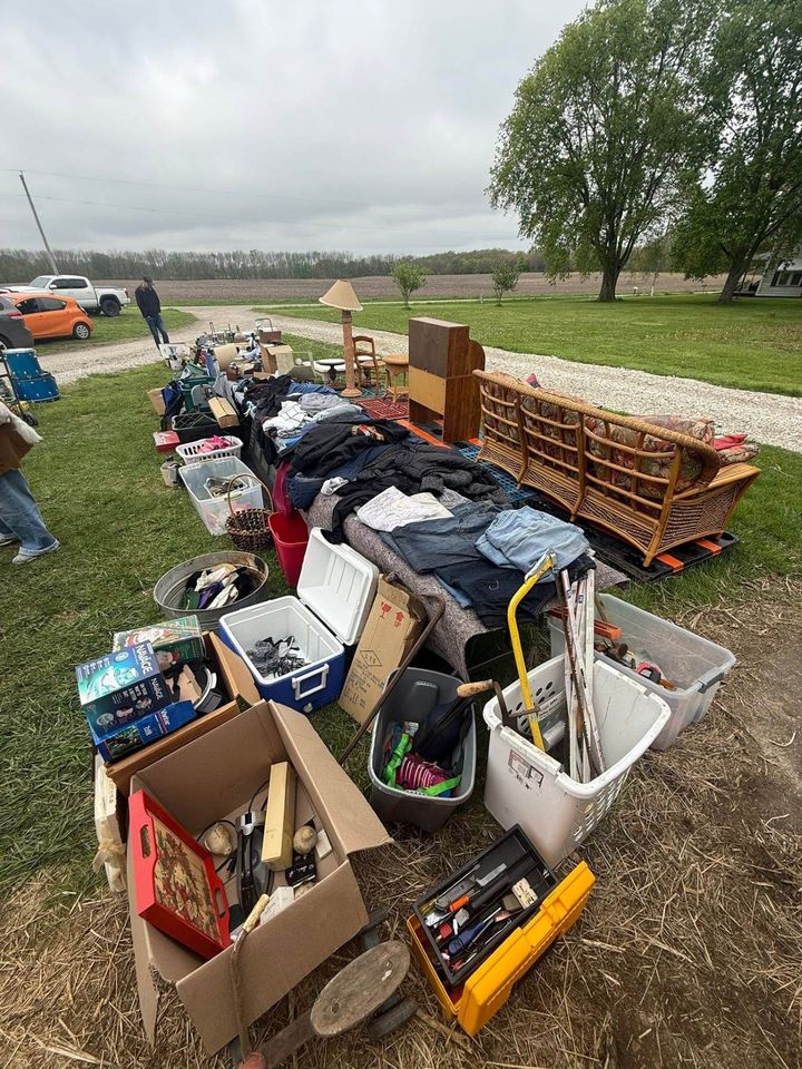 Outdoor flea market table with boxes of assorted clothes and household items on a grassy field under cloudy sky