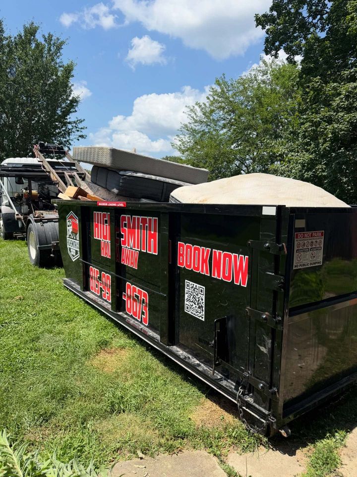Black dumpster filled with sand and debris at a grassy outdoor site under a blue sky