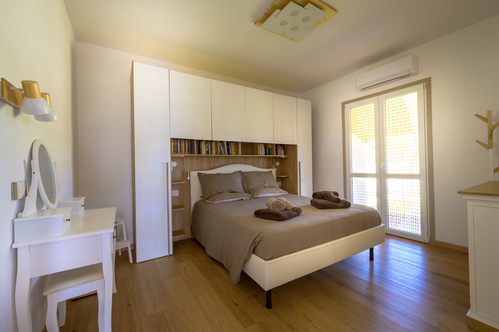 Camera da letto con letto, armadi a muro, mobiletto per il trucco e finestra luminosa. Colori neutri e tenui e pavimenti in legno.