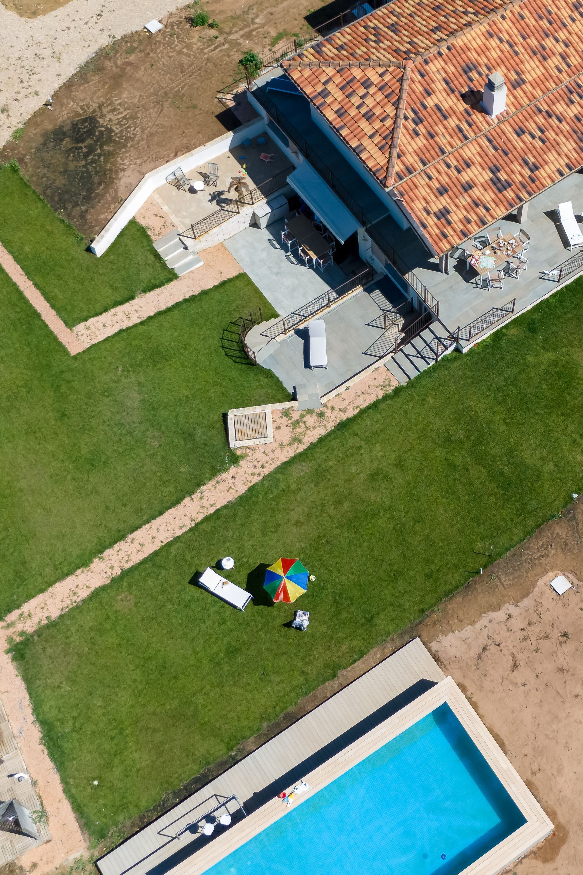 Vista aerea di una casa con tetto di tegole rosse, prato verde, piscina e ombrellone.