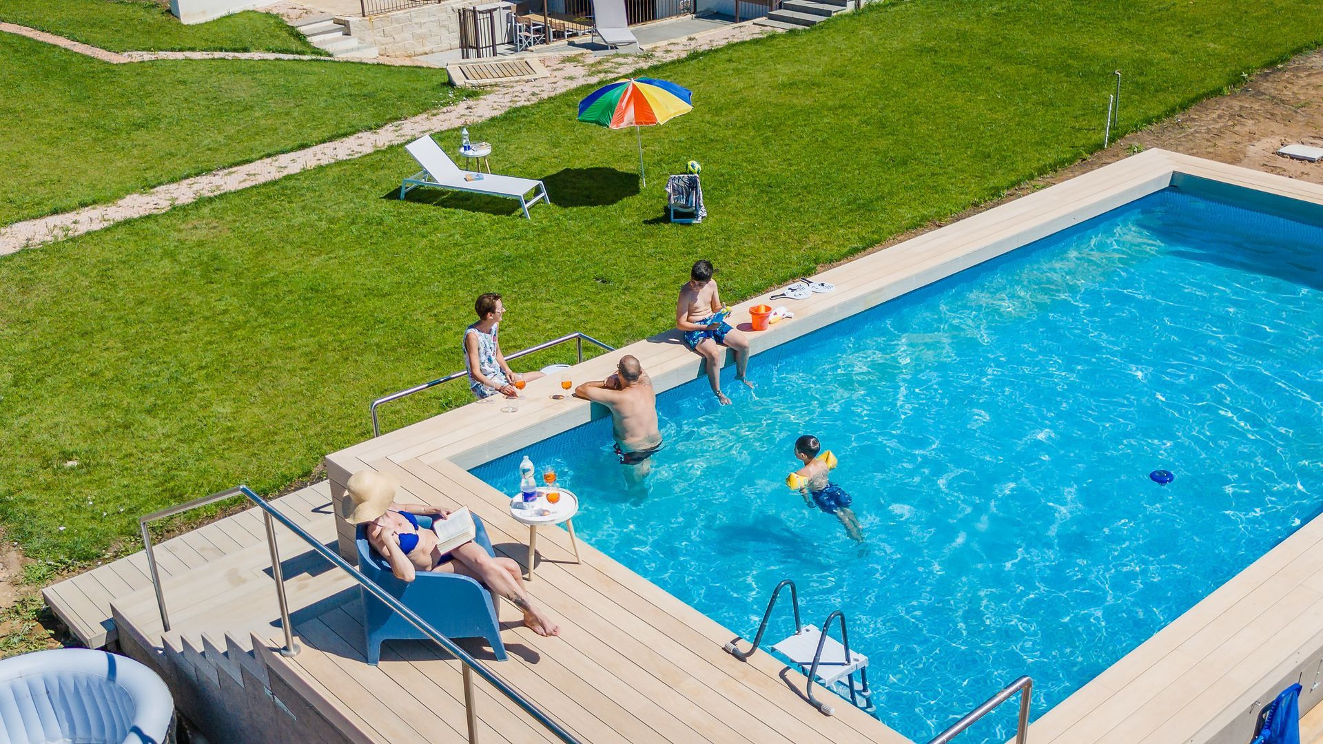 Persone che si rilassano a bordo piscina: prendono il sole, nuotano e sono sedute sul bordo, circondate da un prato verde.