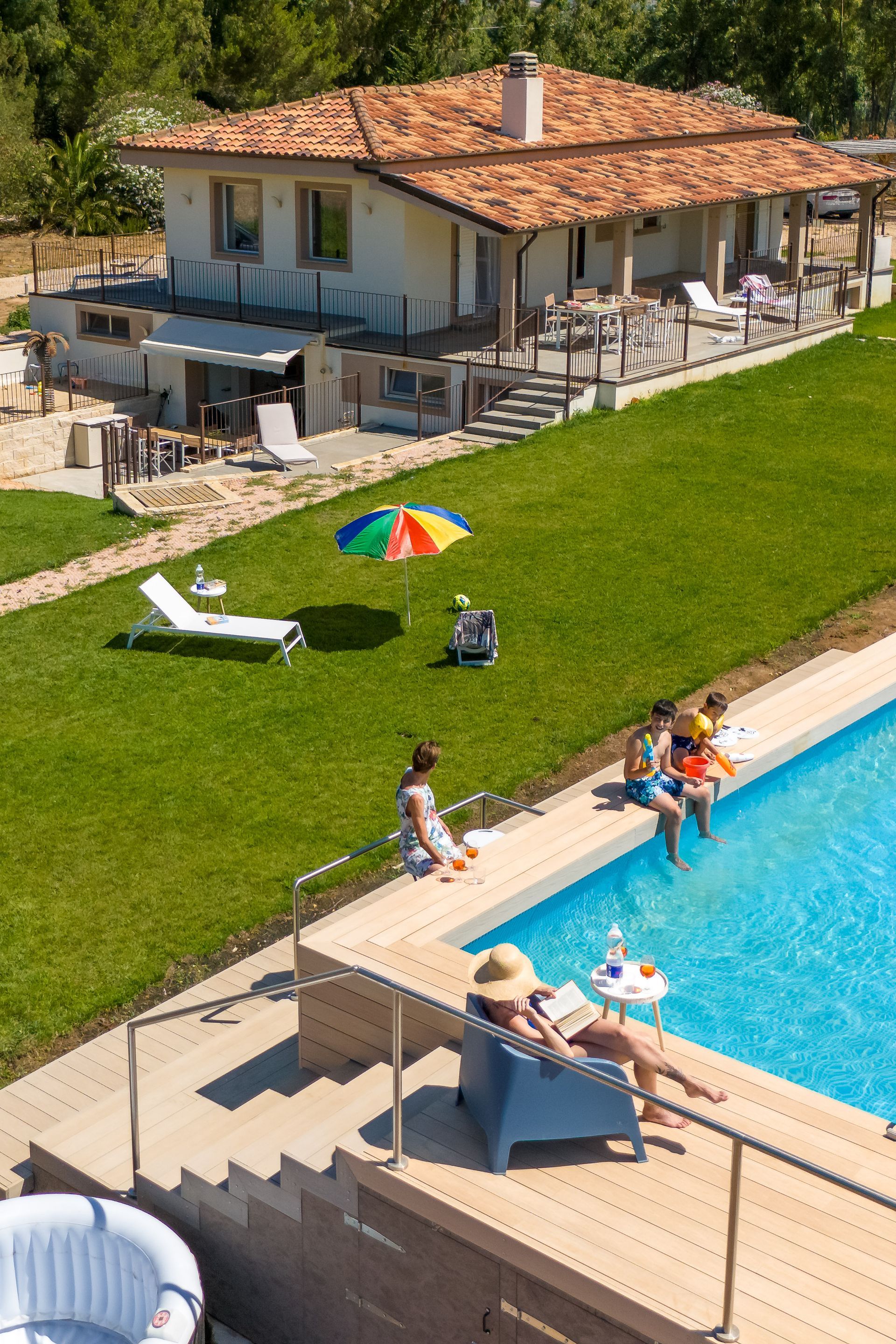 Casa con piscina. Persone che si rilassano vicino all'acqua. Prato verde. Ombrellone, sedie a sdraio.