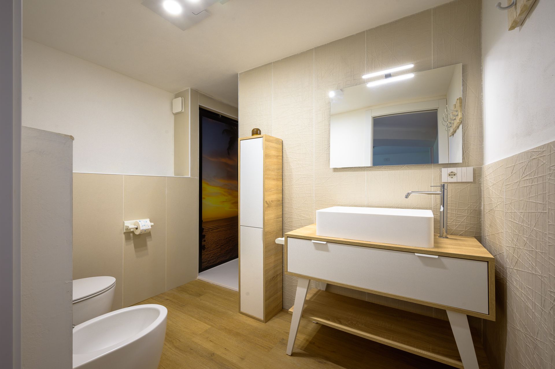 Bagno moderno con lavandino bianco, mobiletto in legno e mobile contenitore alto.