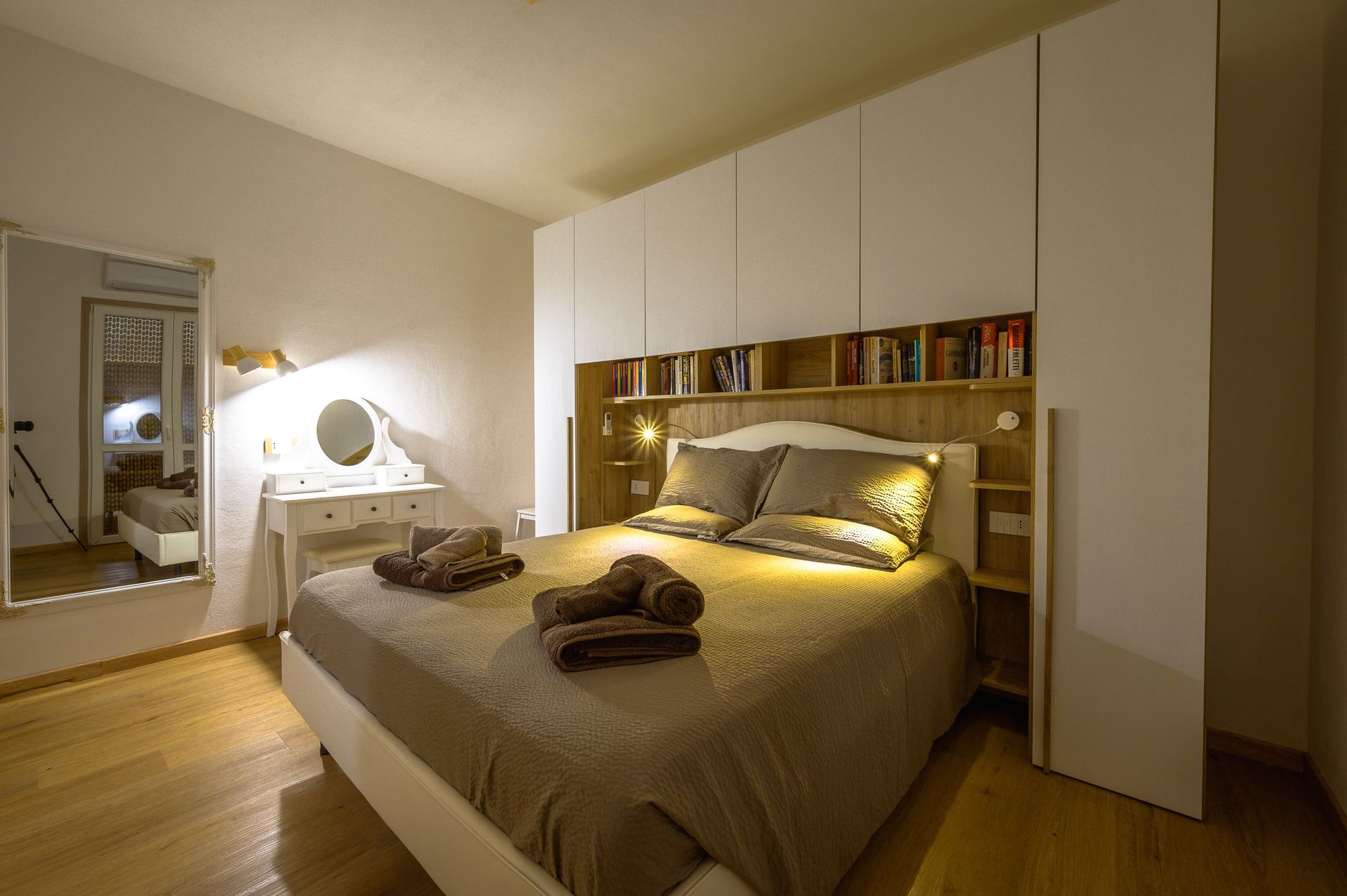 Camera da letto con letto, mobiletto, specchio e armadietti bianchi a muro. Luci soffuse, toni caldi.