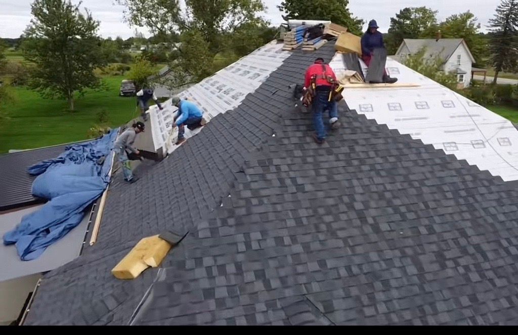 Roofers installing gray shingles on a steep house roof, with white underlayment exposed on the right side.