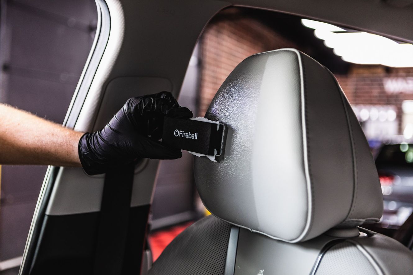 Gloved hand using a tool to clean a car headrest. Interior shot with a white car seat and brick background.