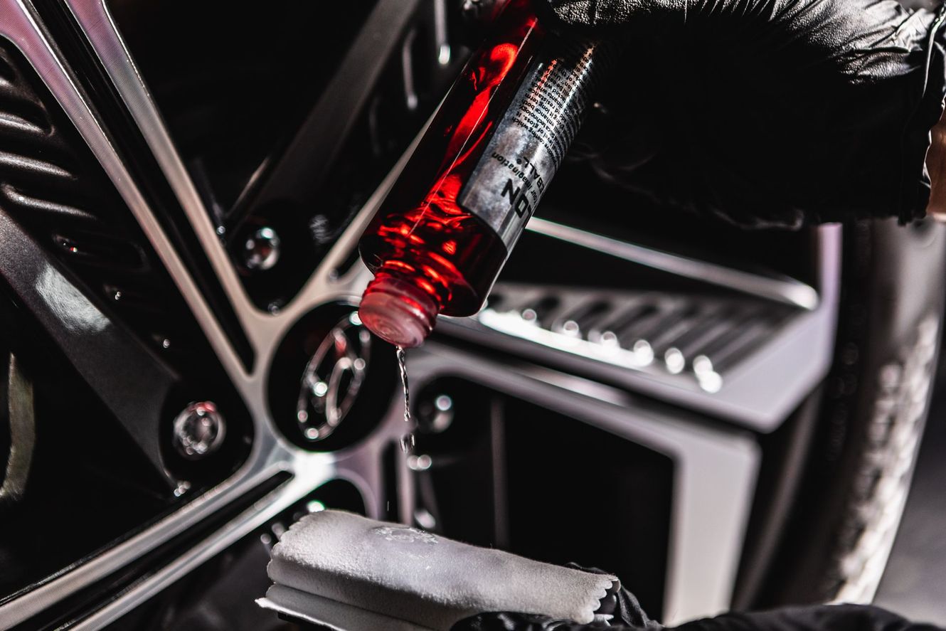 Black-gloved hands applying red liquid to a car wheel with a white applicator.