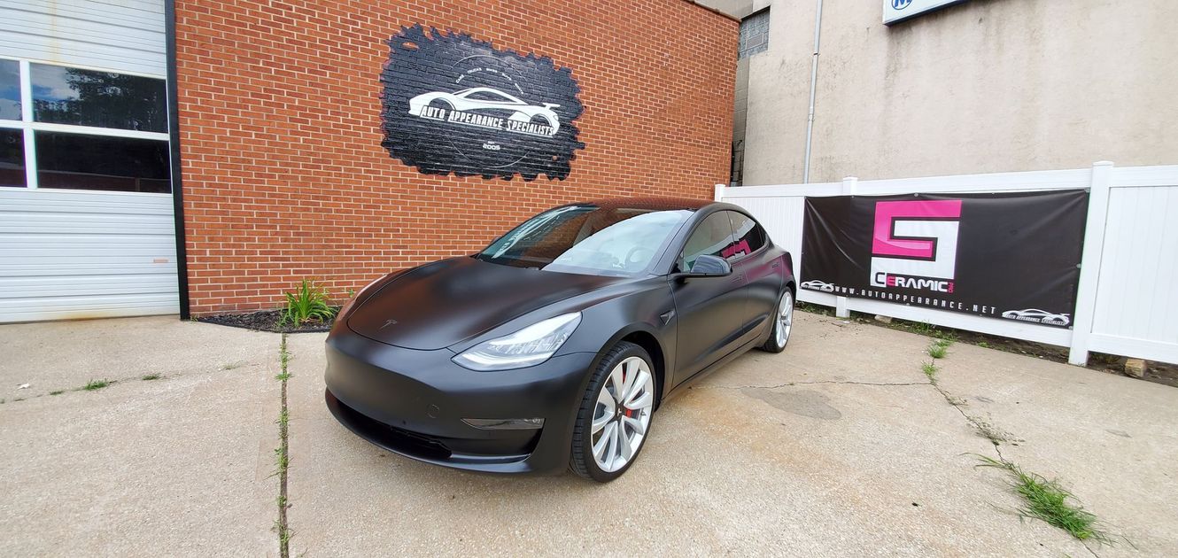 A dark gray Tesla car parked outside of a building. There are signs with logos and a brick wall.