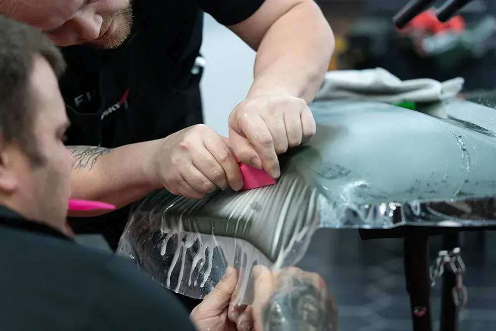 Two people applying film to a car surface with squeegees in a workshop.