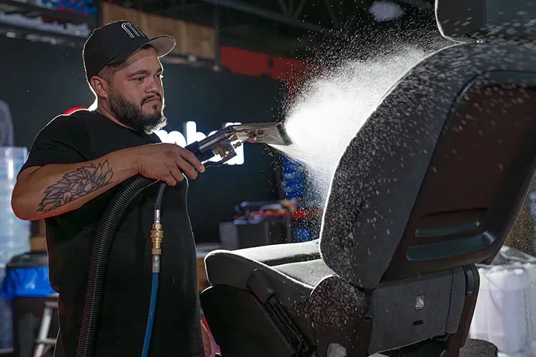 Man uses a cleaning tool on a car seat, spraying white debris. Indoor setting, holding tool in his right hand.