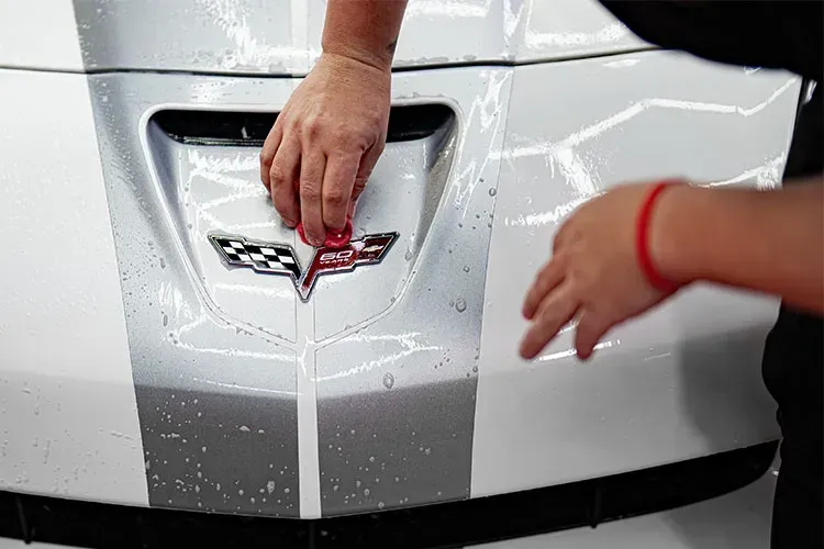 Hands cleaning a white Corvette hood with the car's logo. Stripes are visible.