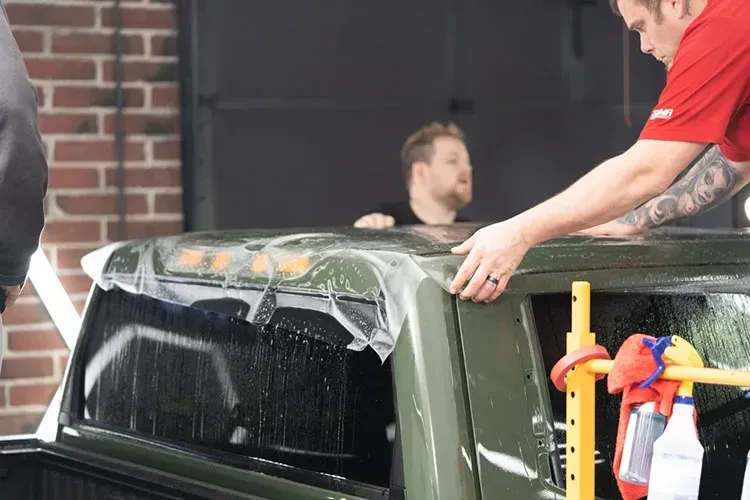 Two people applying tint film to a green truck's windows. A man wears a red shirt and a man wears a black shirt.