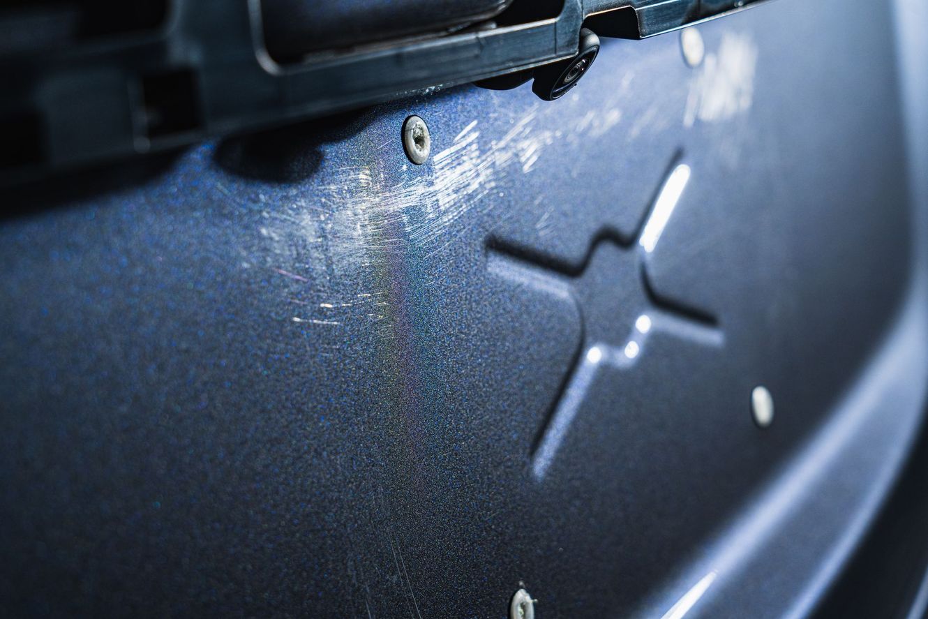 Scratched dark blue car bumper with an X-shaped logo, under a metal plate.