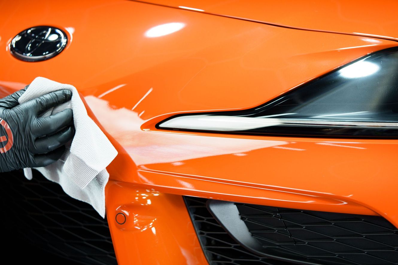 Gloved hand wiping a shiny orange car's front fender with a white cloth.