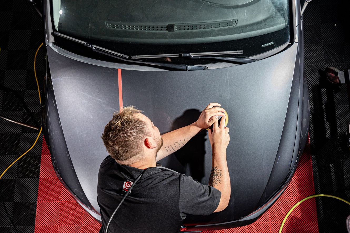 Man polishing a car hood; gray vehicle, dark shirt, indoor setting, red and black tile flooring.