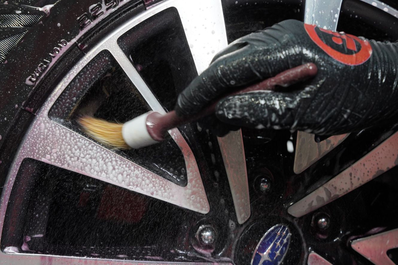 A person wearing a black glove cleans a car rim with a brush; the rim has silver and black detailing.