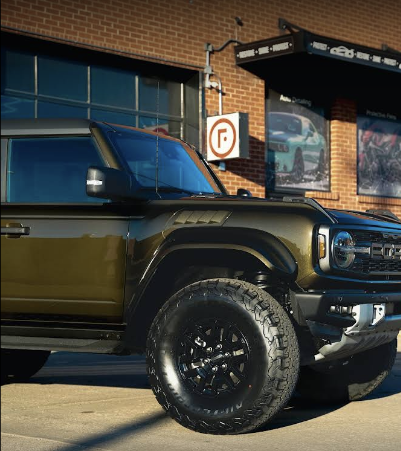 Dark green Ford Bronco with black tires parked in front of a brick building.