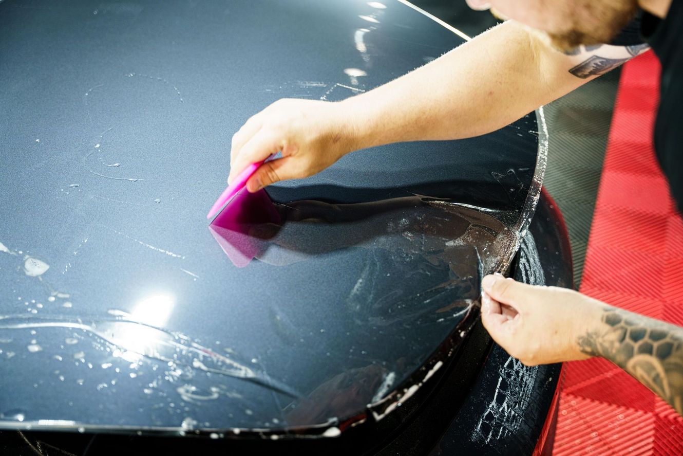 Person applying protective film to a car's hood with a squeegee tool, indoor setting.