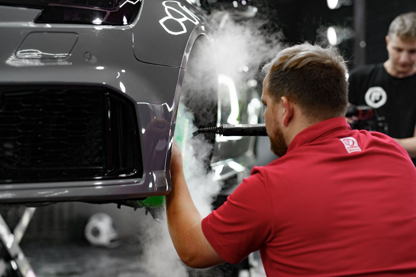 Two men detailing a gray car's bumper, one spraying steam, in a garage.