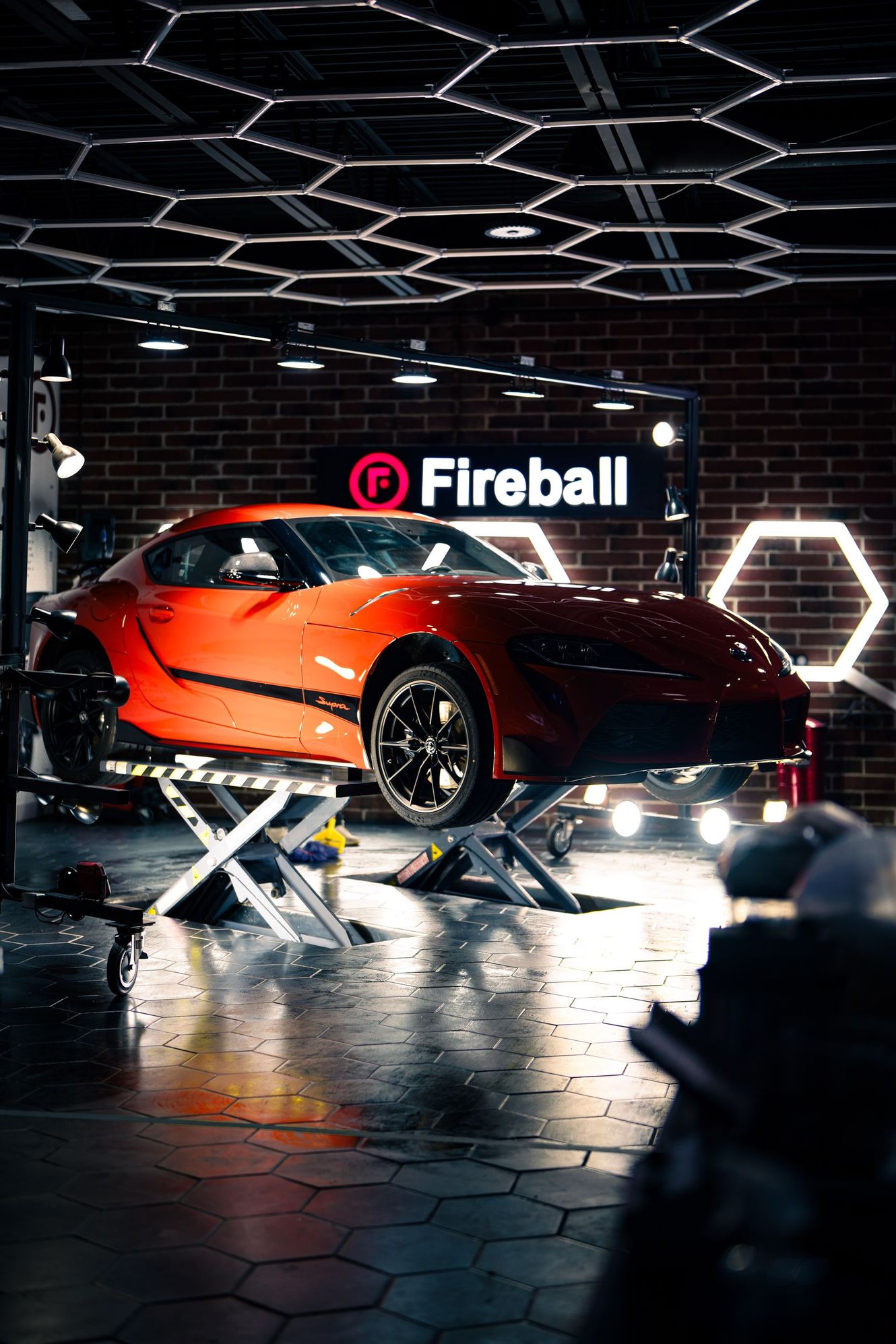Orange sports car on a lift in a garage with hexagonal lights and a Fireball sign.