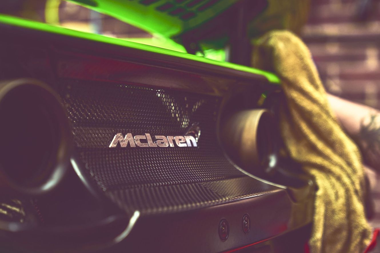 McLaren car rear with logo, exhaust pipe, and someone cleaning with a yellow cloth; close up shot.