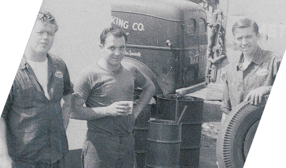 Three men stand beside a large truck and stacked barrels, posing outdoors. | Hunts Point Service Station