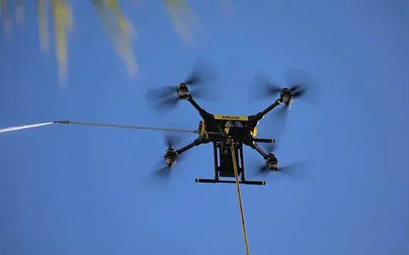 Drone with six rotors flying against a bright blue sky, trailing a line.