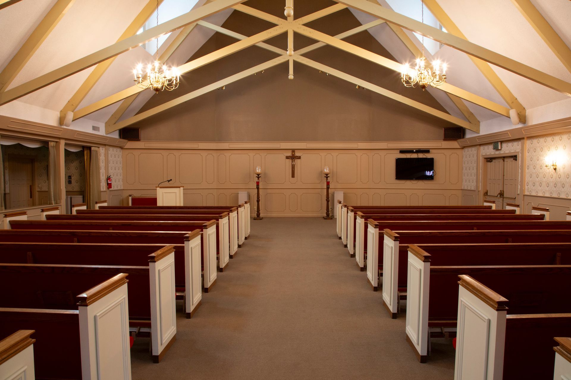 Interior of a chapel with rows of red-cushioned pews facing an elevated coffin, cross, and flowers.