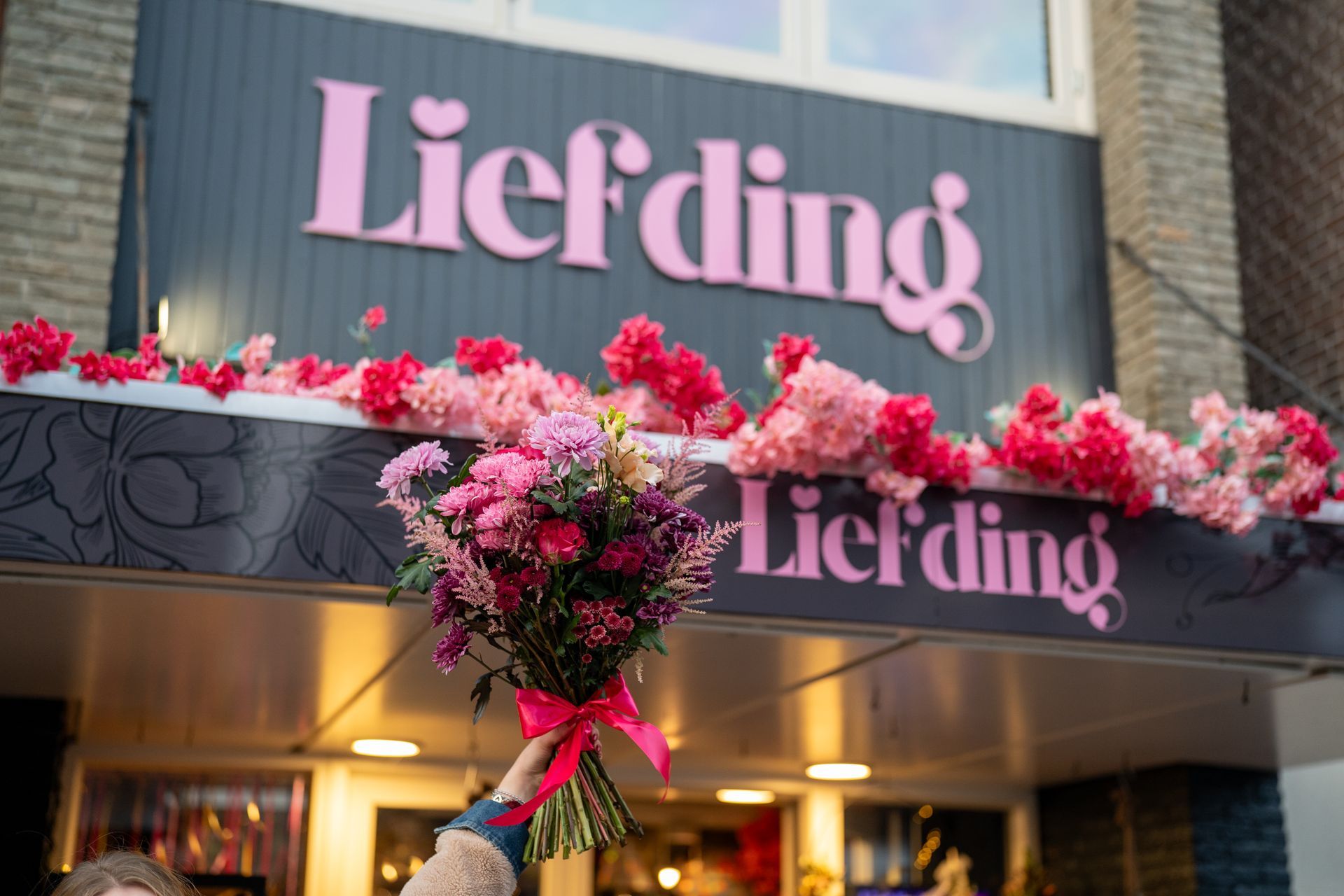 Een persoon met een boeket bloemen met een roze lint staat voor een winkelpui met het roze bordje 