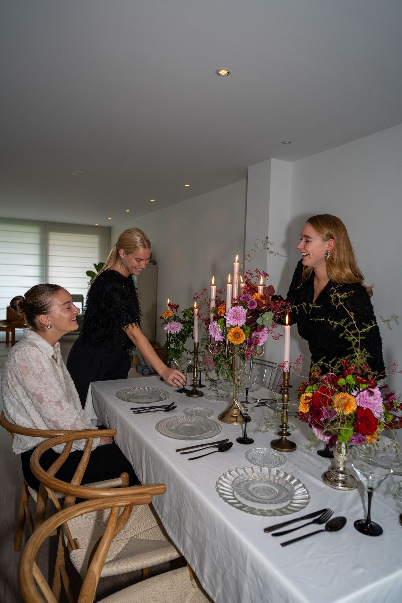 Drie mensen dekken een eettafel die is versierd met bloemstukken, kaarsen en servies in een witte kamer.
