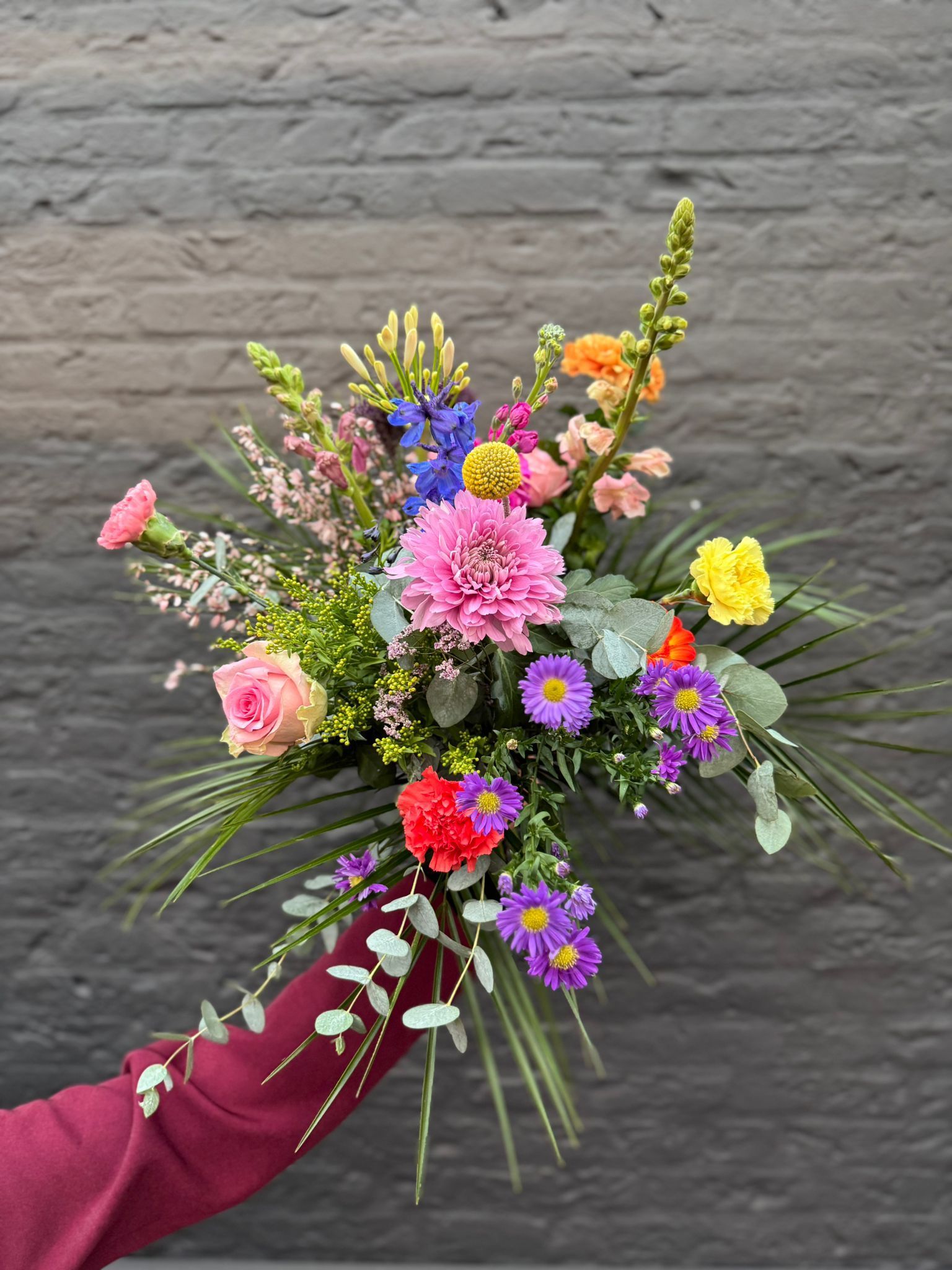 Een hand in een bordeauxrode mouw houdt een levendig, veelkleurig boeket wilde bloemen vast tegen een donkere bakstenen muur.