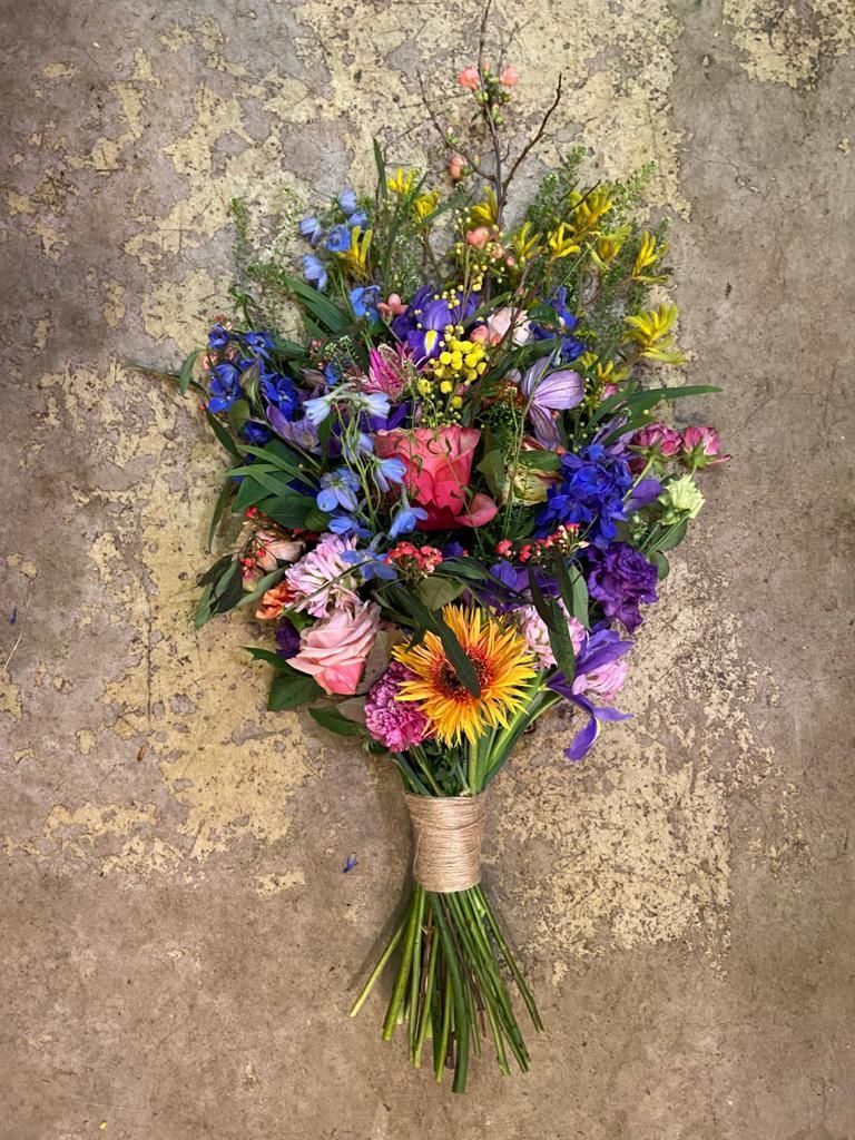Een boeket wilde bloemen met blauwe, roze en gele bloemen, samengebonden met touw, tegen een lichtbruine, gestructureerde achtergrond.