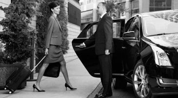 Woman in a suit walking toward a man beside an open black car door outside a building.
