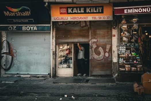 Small row of closed storefronts with signs, a lit shop doorway, and a person standing in the entrance.