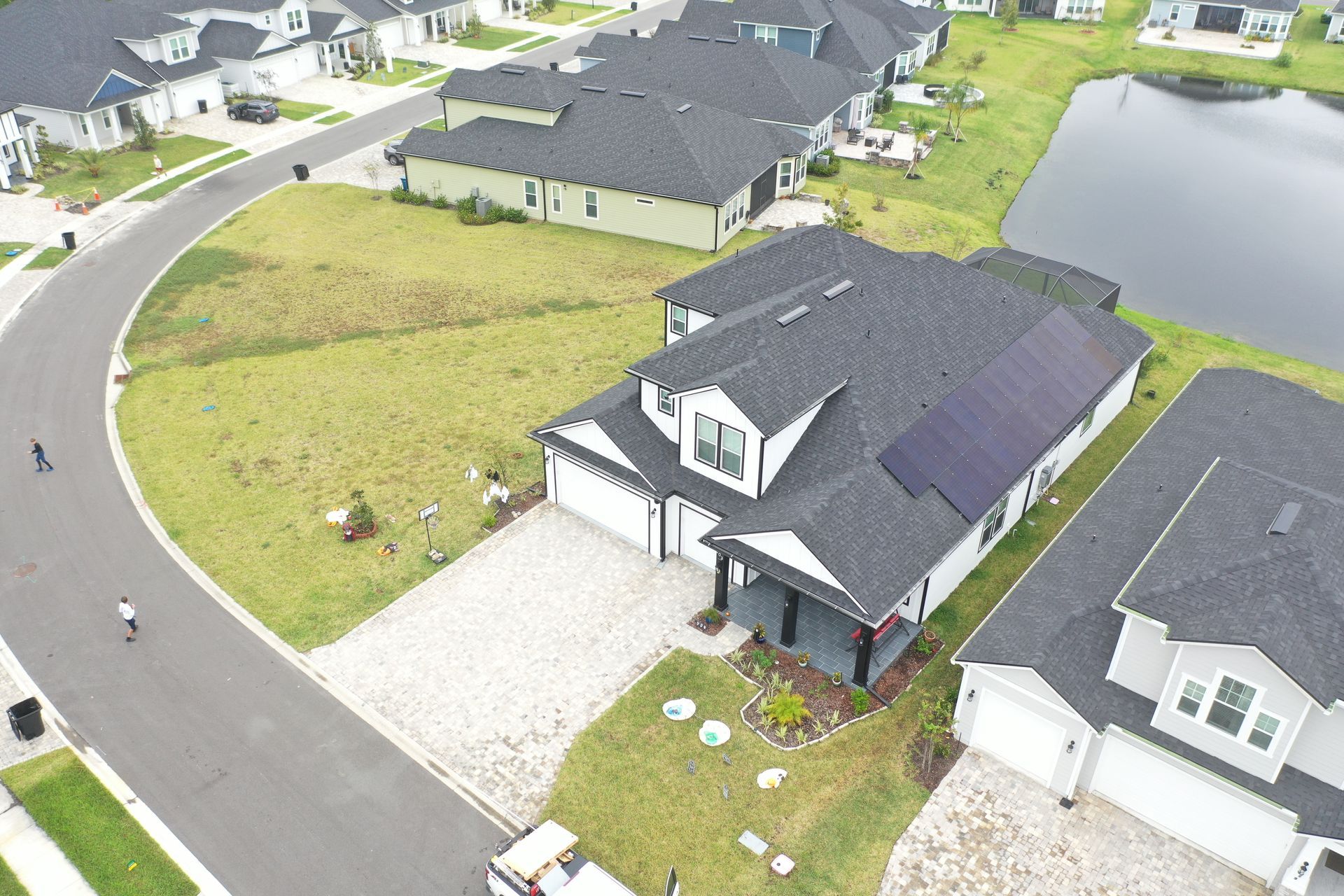 Aerial view of suburban homes with dark roofs, driveways, green lawns, and a pond beside the street.