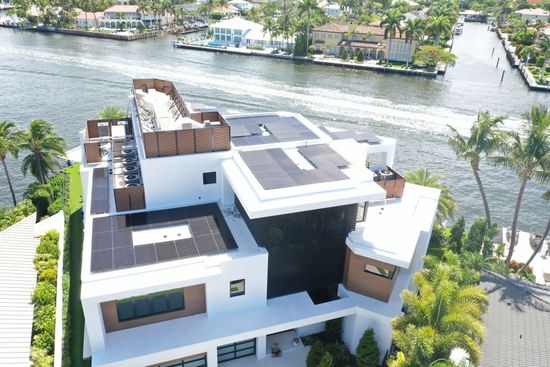 Aerial view of a modern waterfront house with white and dark roofs beside a canal and palm trees.