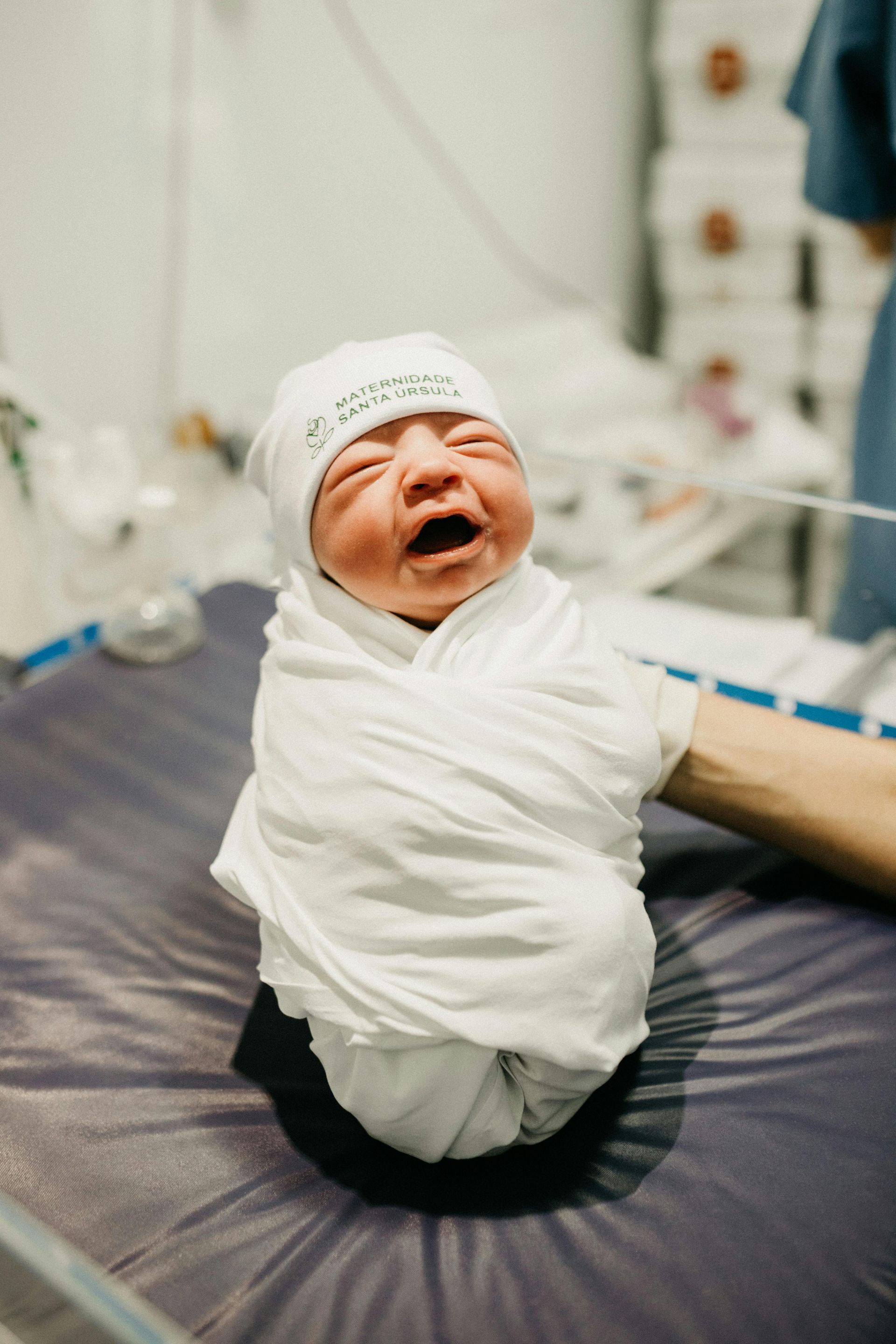 A newborn baby wrapped in a white blanket is crying in a hospital bed.