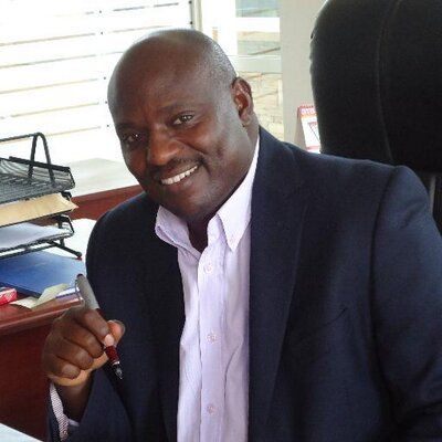 Smiling Black man in a blazer, holding a pen. Sitting at a desk in an office setting.