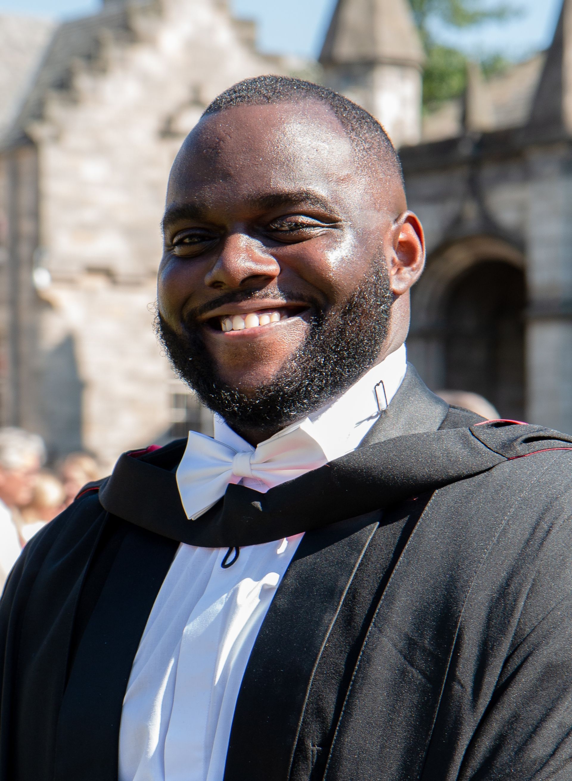 Man in graduation gown smiles broadly outdoors.