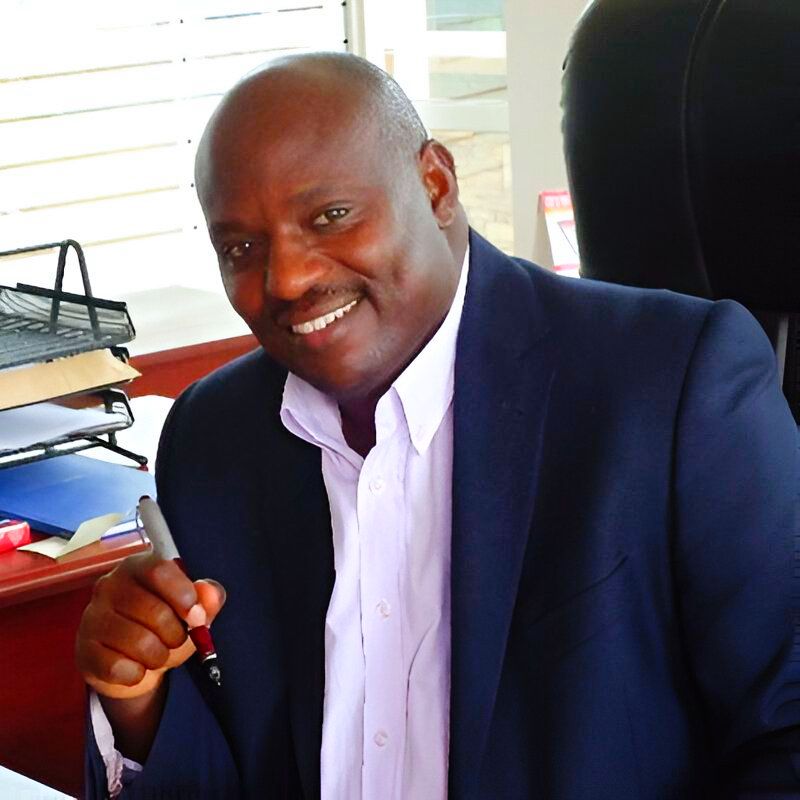 Smiling Black man in a blue blazer at a desk, holding a pen.