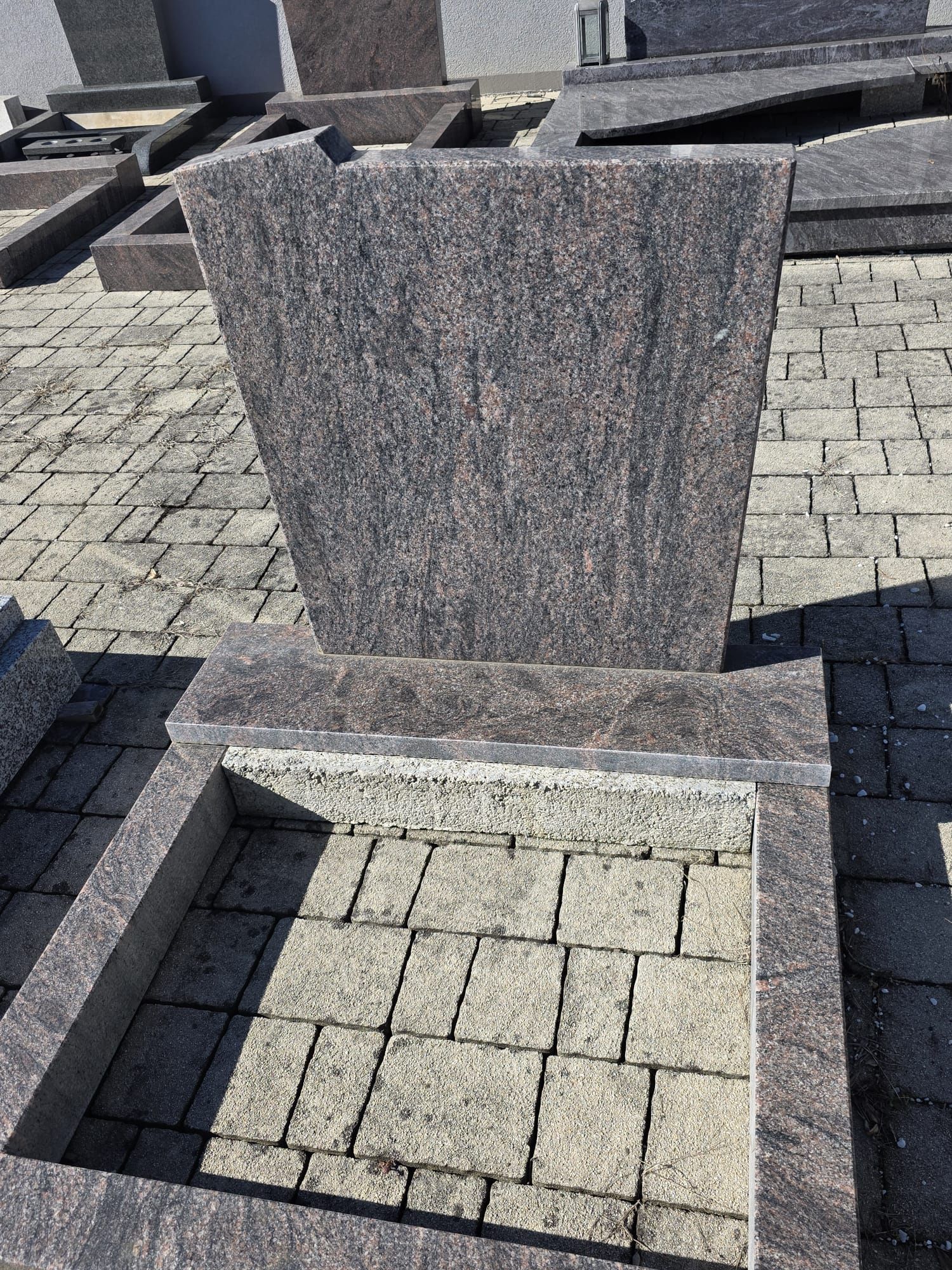 Grabstein auf einem gepflasterten Friedhofsplatz mit Granitumrandung und grauem Steinmonument